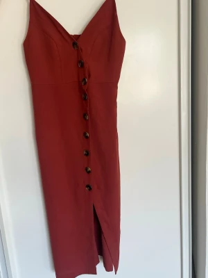 Röd långklänning med knappar - Snygg röd långklänning med smala axelband och knappar framtill. Klänningen har en slits nedtill och figurnära passform. Perfekt för dig som gillar stilrena detaljer och en klassisk look.