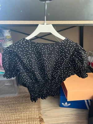 Svart prickig blus från SHEIN - Snygg svart blus från SHEIN med vita prickar, puffärmar och smockad midja. Blusen har v-ringning och knyt framtill, samt korta ärmar med volangkant. Perfekt för dig som gillar trendiga och feminina toppar.