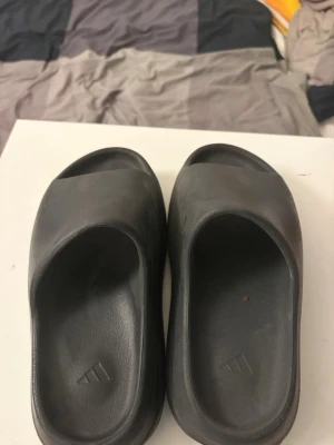 Svarta Adidas tofflor Yeezy Slide - Säljer ett par svarta Adidas Yeezy Slide tofflor med chunky sula och minimalistisk design. Tillverkade i mjukt och flexibelt syntetmaterial med diskret Adidas-logga i fotbädden. Perfekta för chill dagar hemma eller när du vill ha en clean look.