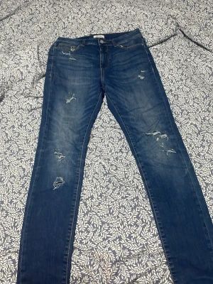Blå slitna skinny jeans - Säljer ett par blå skinny jeans med slitningar och hål på benen. Jeansen har klassisk femficksdesign, låg midja och smal passform. Materialet är denim med stretch för extra komfort. Perfekta för en avslappnad och trendig look.