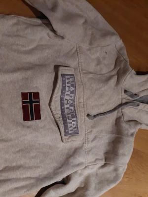 Grå hoodie från Napapijri med flagga - Säljer en grå hoodie från Napapijri med stor logga och norsk flagga på bröstet. Hoodien har huva med dragsko och en stor magficka framtill. Tillverkad i mjukt sweatshirtmaterial, perfekt för chill dagar.