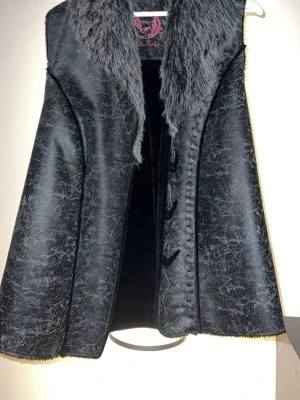 Svart pälsväst med fuskpälsdetaljer - Stilren svart pälsväst med bred krage i mjuk fuskpäls och dekorativa knappar framtill. Västen har en lätt glansig yta och markerade sömmar som ger en cool vintagekänsla. Perfekt för dig som vill addera lite edge till din outfit.