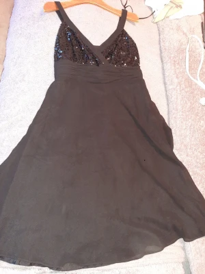 Svart paljettklänning med v-ringning från Debenhams - Snygg svart klänning med breda axelband och v-ringning. Överdelen är dekorerad med svarta paljetter som ger ett glittrigt intryck. Kjolen är lätt utsvängd och klänningen är kort i modellen. Perfekt för dig som vill sticka ut lite extra.