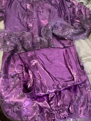 Kurdisk newroz klänning med byxa - Unik lila långklänning med transparent spets och blommiga paljettdetaljer. Klänningen har volangdetaljer och ett glansigt innerfoder som ger extra lyxig känsla. Perfekt för dig som vill sticka ut med en elegant och färgstark look.