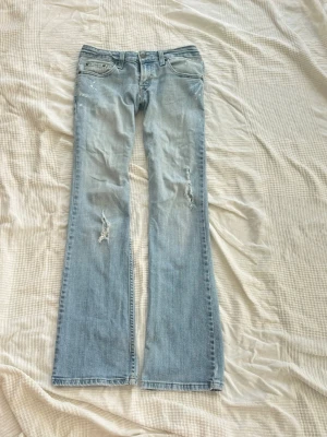Ljusblå lowwaist bootcut jeans med slitningar - Säljer ett par ljusblå bootcut jeans med slitningar på båda benen. Jeansen har klassisk femficksmodell och låg midja. Perfekta för dig som gillar en avslappnad och trendig stil. Materialet är denim i bomull.
