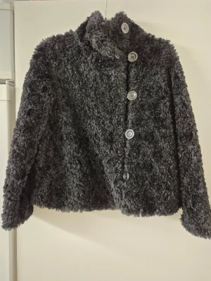 Teddyjacka - Mysig svart teddyjacka med hög krage och fyra stora knappar framtill. Jackan har en fluffig, mjuk struktur och är tillverkad i syntetmaterial som ger en riktigt gosig känsla. Perfekt för kyliga dagar när du vill vara både varm och snygg.