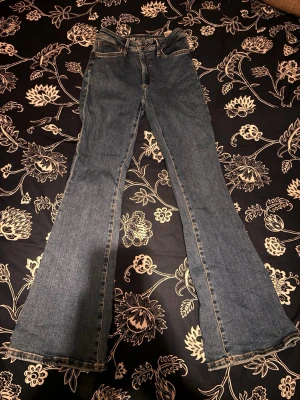 Blå bootcut jeans med kontrastsömmar - Säljer ett par klassiska blå bootcut jeans med kontrastsömmar och femficksmodell. Jeansen har normal midja och utsvängda ben, perfekta för en retroinspirerad look. Materialet är denim i bomull och de har bälteshällor samt dragkedjegylf.