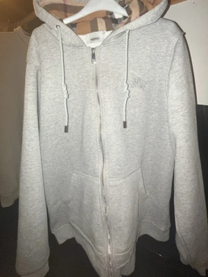 Grå zip-hoodie från Burberry - Snygg grå hoodie med dragkedja från Burberry. Klassisk huva med justerbara snören och ikoniskt rutigt foder. Mjuk bomullsmix, två fickor fram och diskret logga på bröstet. Perfekt för en chill och stilren look.