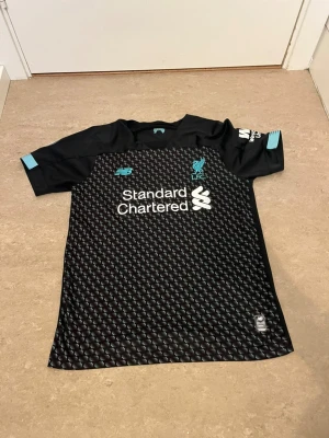 Liverpool bortatröja Fabinho #3 - Snygg svart Liverpool FC bortatröja från New Balance med turkosa detaljer och mönstrad framsida. Tryck med Fabinho och nummer 3 på ryggen. Officiella Premier League-märken på ärmen och Standard Chartered som sponsor på bröstet. Perfekt för fotbollsfans!