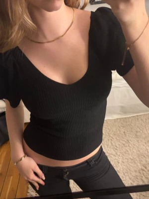 Svart ribbad croppad topp - Snygg svart croppad topp med ribbad struktur och v-ringning. Toppen har korta puffärmar och sitter tight mot kroppen för en modern look. Ryggen har band som går att knyta och toppen är i väldigt bra skick.