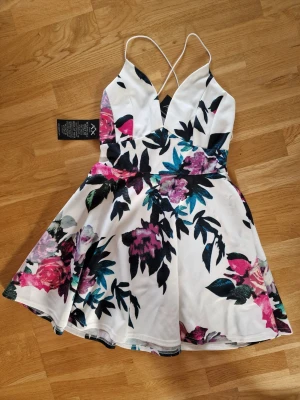 Vit blommig kortklänning AX Paris - Supersöt vit kortklänning från AX Paris med färgglatt blommönster i rosa, lila och blå toner. Klänningen har smala axelband som korsas i ryggen, djup v-ringning och en utställd kjol. Perfekt för sommarens alla tillfällen!
