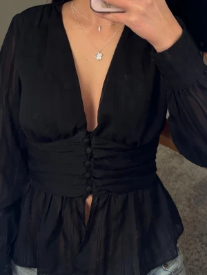 Svart blus med markerad midja - Stilren svart blus med djup v-ringning och markerad midja med knappar framtill. Blusen har långa, lätt transparenta ärmar och ett tunt, skönt material som ger en elegant look. Perfekt att matcha med jeans eller kjol för en trendig outfit.