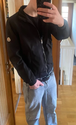 Svart Moncler vindjacka med dragkedja S - Snygg svart vindjacka från Moncler med klassisk logga på ärmen. Jackan har hel dragkedja framtill, två fickor och ribbade muddar vid ärmslut och midja. Perfekt nu till våren för en stilren och sportig look.