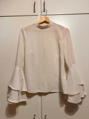 Vit blus med vida ärmar - Jättefin blus med volangärmar. I bra skick utöver att det finns en liten fläck på ena ärmen som inte gått bort i tvätten. Se sista bilden. Fläcken stör inte alls mycket då den knappt syns när man har på sig toppen.