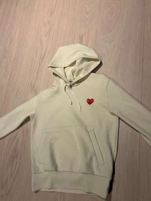 Gul hoodie från Comme des Garçons - Snygg ljusgul hoodie från Comme des Garçons med det ikoniska röda hjärtat på bröstet. Hoodien har huva med dragsko, stor magficka och ribbade muddar. Perfekt för dig som vill ha en clean look med en twist. Materialet känns mjukt och skönt mot huden.