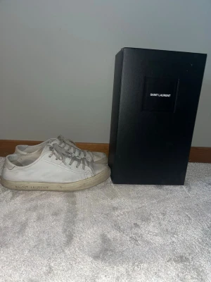 Vita Saint Laurent sneakers i läder - Säljer ett par klassiska vita sneakers från Saint Laurent i läder med diskret logga på sidan. Skorna har lågt skaft, vita snören och en stilren design som passar till det mesta. Kommer med originalkartong. Skorna har ett litet hål på vänster sko på sidan, men inget som läggs märke till. Se slide 5 för bild.