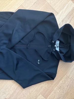Svart hoodie från Comme des Garçons Play - Svart hoodie från Comme des Garçons Play med dragkedja framtill och justerbar huva. På bröstet sitter det ikoniska svarta hjärtat med ögon. Tillverkad i mjuk bomull, perfekt för en avslappnad och stilren look.