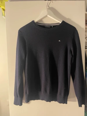 Mörkblå stickad tröja J.Lindeberg - Mörkblå stickad tröja från J.Lindeberg med rund halsringning och diskret logga på bröstet. Tröjan har långa ärmar och ribbade muddar vid ärmslut och nederkant. Perfekt för lager-på-lager och stilrena outfits.