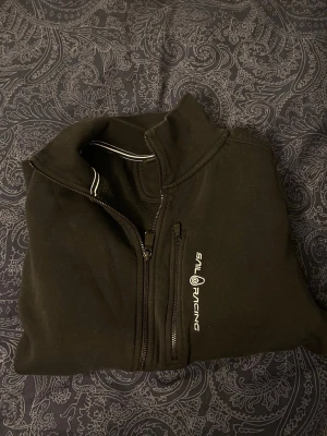 Svart Sail Racing ziptröja M - Säljer en svart ziptröja från Sail Racing med halv dragkedja och vit logga på bröstet. Tröjan har hög krage och diskreta vita detaljer vid kragen. Perfekt för dig som gillar sportig och stilren look.