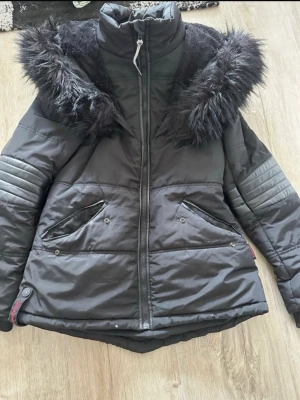 Svart pufferjacka med fuskpäls - Snygg svart pufferjacka med hög krage och stor huva med svart fuskpäls. Jackan har quiltade detaljer på ärmarna, två snedställda fickor med tryckknappar och dragkedja framtill. Perfekt för kalla vinterdagar och riktigt varm.