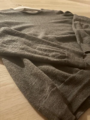 Mörkgrå merinoulltröja från H&M - Snygg mörkgrå stickad tröja från H&M i mjuk merinoull. Tröjan har rund halsringning, långa ärmar och ribbade muddar vid ärmslut och nederkant. Perfekt för lager på lager och enkel att matcha med olika outfits.