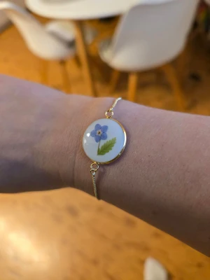 Armband med förgätmigej och blad - Unikt armband med en rund vit platta i mitten, dekorerad med en blomma och ett grönt blad. Plattan är infattad i guldfärgad metall och armbandet har en tunn kedja. Perfekt för dig som gillar naturinspirerade accessoarer.