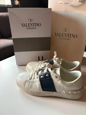 Valentino Garavani vita sneakers - Snygga vita sneakers från Valentino Garavani med marinblå detalj och nitar på hälen. Skorna har lågt skaft, vita snören och är tillverkade i läder. Perfekta för dig som vill ha en exklusiv och stilren look.