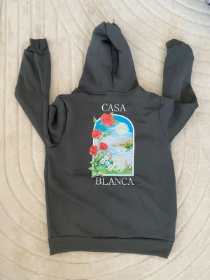 Casa Blanca hoodie med rosor - Mörkgrå hoodie från Casa Blanca med stor färgglad ryggtryck av rosor och landskap i fönsterdesign. Framsidan har samma motiv i mindre format och texten 'Casa Blanca'. Utrustad med huva och känguruficka. Perfekt för en chill och trendig look: Storleken kan vara S eller M.