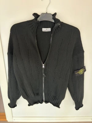 Svart stickad ziptröja Stone Island - Säljer en svart stickad ziptröja från Stone Island med ikonisk patch på ärmen. Tröjan har hög krage, ribbade muddar och dragkedja framtill. Materialet är grovstickat och ger en schysst streetwear-vibe.