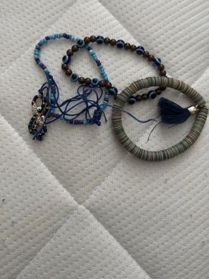 Färgglada armband med pärlor och tofs - Säljer ett set med armband i blåa och bruna toner. Ena armbandet har små pärlor i olika nyanser av blått och brunt, samt ögonmönster. Det andra är ett grönt-randigt armband med blå tofs. Perfekt för dig som gillar boho-stil och unika accessoarer.