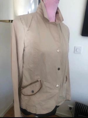 Beige jacka med hög krage - Stilren beige jacka med hög krage och knappstängning framtill. Jackan har två snedställda fickor med dragkedja och knappdetalj samt markerad midja baktill för en snygg siluett. Tillverkad i ett lätt material som passar perfekt till vår och höst.