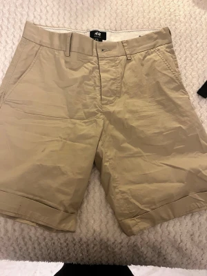 Beige shorts från H&M med fickor - Snygga beige shorts från H&M med klassisk passform. Shortsen har bälteshällor, sidofickor och två bakfickor med knapp. Tillverkade i ett lätt bomullsmaterial, perfekta för varma dagar. Stängs med dragkedja och knapp framtill. Obs: herr shorts.