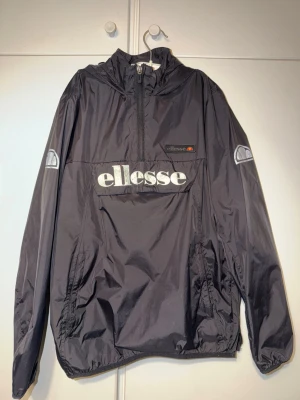 Svart vindjacka från Ellesse - Svart vindjacka från Ellesse med stor logga framtill, halv dragkedja och huva. Jackan har en stor ficka på magen och reflexdetaljer på ärmarna. Tillverkad i lätt och glansigt material, perfekt för en sportig och avslappnad stil.