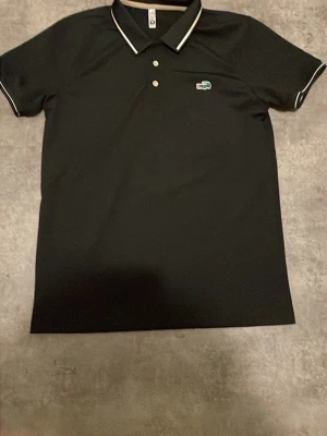 Svart pikétröja från Lacoste - Klassisk svart pikétröja från Lacoste med vit kant på kragen och ärmsluten. Tröjan har tre knappar framtill och den ikoniska gröna krokodilloggan på bröstet. Perfekt för en stilren och sportig look.