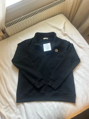 Svart sweatshirt från Moncler - Snygg svart sweatshirt från Moncler med halv dragkedja och klassisk logga på bröstet. Tröjan har ribbade muddar vid ärmslut och nederkant samt en hög krage. Tillverkad i mjukt bomullsmaterial som känns skönt mot huden. Perfekt för en clean och stilren look.