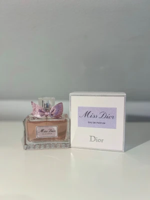 Miss Dior Eau de Parfum från Dior - Miss Dior Eau de Parfum från Dior i en elegant glasflaska med ljusrosa doft och en söt, glittrig rosett runt halsen. Flaskan har en fyrkantig form och kommer med originalkartong i vitt och lila. Perfekt för dig som älskar lyxiga och feminina parfymer.