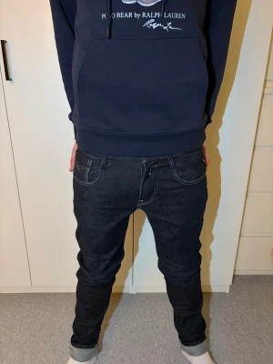 Svarta jeans från replay - Säljer ett par svarta jeans från Replay med klassisk kontrastsöm och unik bakficka med broderad logga och liten röd detalj. Jeansen har en slim fit passform och är tillverkade i kraftigt denimtyg. Perfekta för dig som gillar streetwear och snygga detaljer. Storlek 29/32