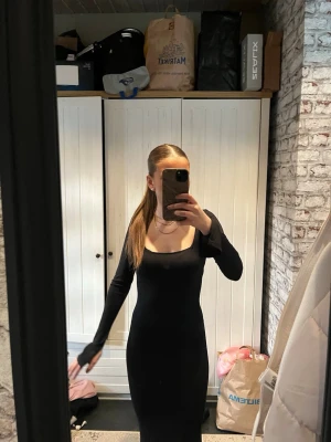 Svart långklänning med lång ärm - En elegant svart långklänning med figurnära passform och långa ärmar. Klänningen har en fyrkantig urringning och är enkel men stilren, perfekt för dig som gillar minimalistisk design. Materialet ser ut att vara mjukt och stretchigt, vilket ger en bekväm känsla.