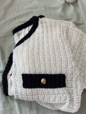 Vit kofta med svarta detaljer och guldknappar - Stilren vit stickad kofta med svarta ribbade kanter och dekorativa fickor framtill. Guldiga knappar ger en lyxig touch. Koftan har rund halsringning och är tillverkad i ett grovstickat material. Perfekt för att lyfta din outfit.