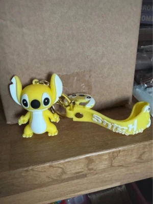 Gul Stitch nyckelring med figur - Söt nyckelring med en gul Stitch-figur i mjukt gummi och matchande band med texten 'Stitch Lovely Baby'. Perfekt accessoar till väskan eller nycklarna för dig som gillar tecknade figurer.