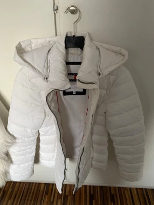 Vit pufferjacka från Tommy Hilfiger - Snygg vit pufferjacka från Tommy Hilfiger med huva och dragkedja framtill. Jackan har diskret logga på bröstet och quiltad design. Perfekt för kalla dagar med sin vadderade och värmande känsla. Figursydd passform och stilren look.