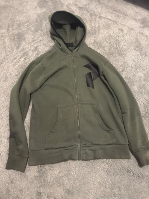 Peak Performance hoodie/kofta - Snygg olivgrön hoodie från Peak Performance med dragkedja framtill och stor ficka på magen. Har en svart logga på bröstet och klassisk huva. Tillverkad i mjukt bomullsmaterial som känns skönt mot huden. Perfekt för en avslappnad och sportig stil.