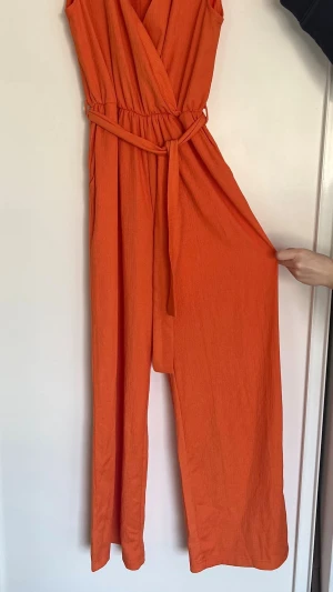 Ärmlös orange jumpsuit med bälte - Snygg ärmlös orange jumpsuit med v-ringning och matchande tygbälte i midjan. Tillverkad i ett mjukt, stretchigt tyg med lätt struktur. Perfekt för dig som vill sticka ut med färg och bekväm passform.
