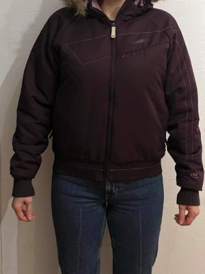 Mörklila pufferjacka från Helly Hansen - Snygg mörklila pufferjacka från Helly Hansen med huva och fuskpälsdetalj. Jackan har dragkedja framtill, ribbade muddar och en bröstficka med dragkedja. Perfekt för kalla vinterdagar och ger en sportig look. Modellen på bilden är 169cm.