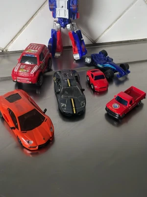 Samling leksaksbilar och robot - Säljer en samling leksaksbilar i olika färger och modeller, inklusive en orange sportbil, svart bil med gula ränder, röd pickup, röd rallybil, blå formelbil och en blå-röd robotfigur. Perfekt för dig som gillar bilar och robotar.