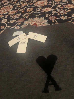 Grå stickad tröja från Ami Paris - Snygg grå stickad tröja från Ami Paris med deras ikoniska svarta hjärta och A-logga på bröstet. Bra kvalité, stickad (bomull/tyg), varm o perfekt för alla årstider.