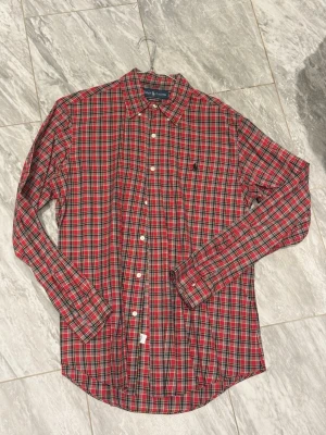 Rutig röd skjorta från Ralph Lauren - Säljer en klassisk rutig skjorta från Ralph Lauren i rött, grönt och gult. Skjortan har lång ärm, knappar framtill och en liten broderad logga på bröstet. Modellen är Custom Fit och passar perfekt till en avslappnad stil.