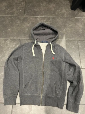 Grå zip hoodie från Polo Ralph Lauren - Mörkgrå hoodie med dragkedja från Polo Ralph Lauren. Klassisk huva med vita snören och röd broderad logga på bröstet. Mjuk bomullsblandning och två fickor framtill. Perfekt för en chill och stilren look.