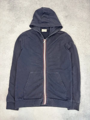 Mörkblå hoodie från Moncler - Snygg mörkblå hoodie från Moncler med dragkedja framtill och klassiska röda, vita och blå detaljer längs dragkedjan. Utrustad med huva och två stora fickor framtill. Perfekt för en avslappnad och stilren look.