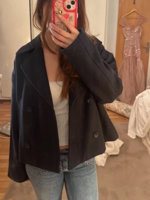 Kort svart dubbelknäppt rock från Selected - Säljer en stilren, kort svart rock från Selected med dubbelknäppning och bred krage. Rocken har raka fickor framtill och är fodrad för extra komfort. Perfekt för dig som gillar en klassisk men trendig look.